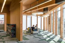 Two men sitting in chairs in front of floor-to-ceiling windows divided by wooden posts. A wooden wall with a bookshelf faces the windows and creates a three-sided space.